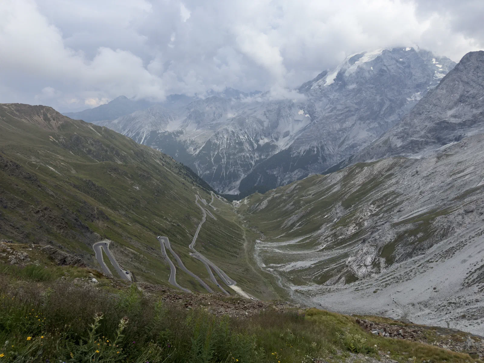 The hairpins of the Stelvio Pass road wind down a green alpine valley, with a glaciated peak under low clouds behind.