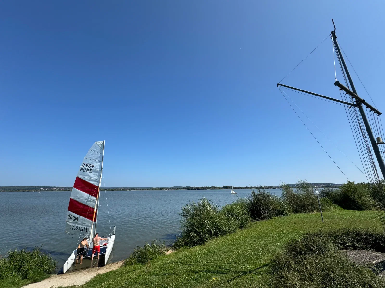 A catamaran with a red-and-white sail on the shore of a lake under a blue sky