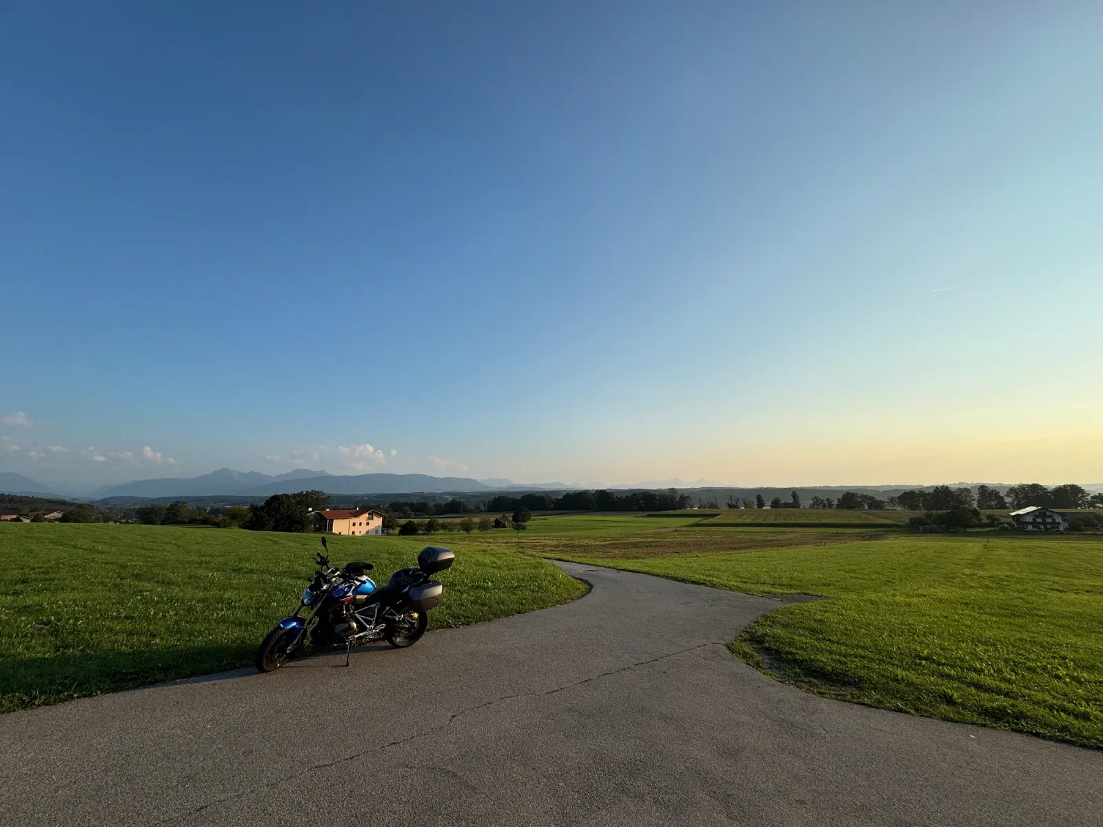 The loaded R 1250 R on a farm road in evening light, the Alps behind it.