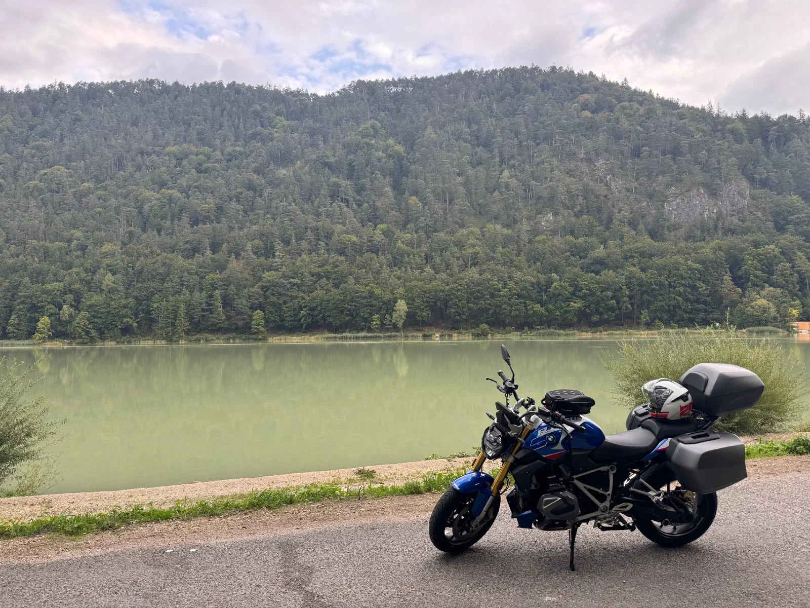 A blue BMW R 1250 R with side cases and a top case stands at the roadside, next to it a greenish lake and a wooded slope, a white helmet lies on the passenger seat