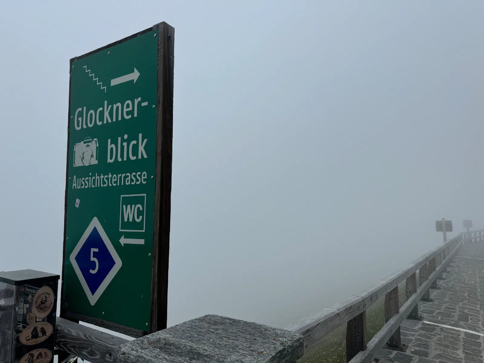 A green sign reading Glocknerblick Aussichtsterrasse, to the right a wooden railing that disappears into thick fog
