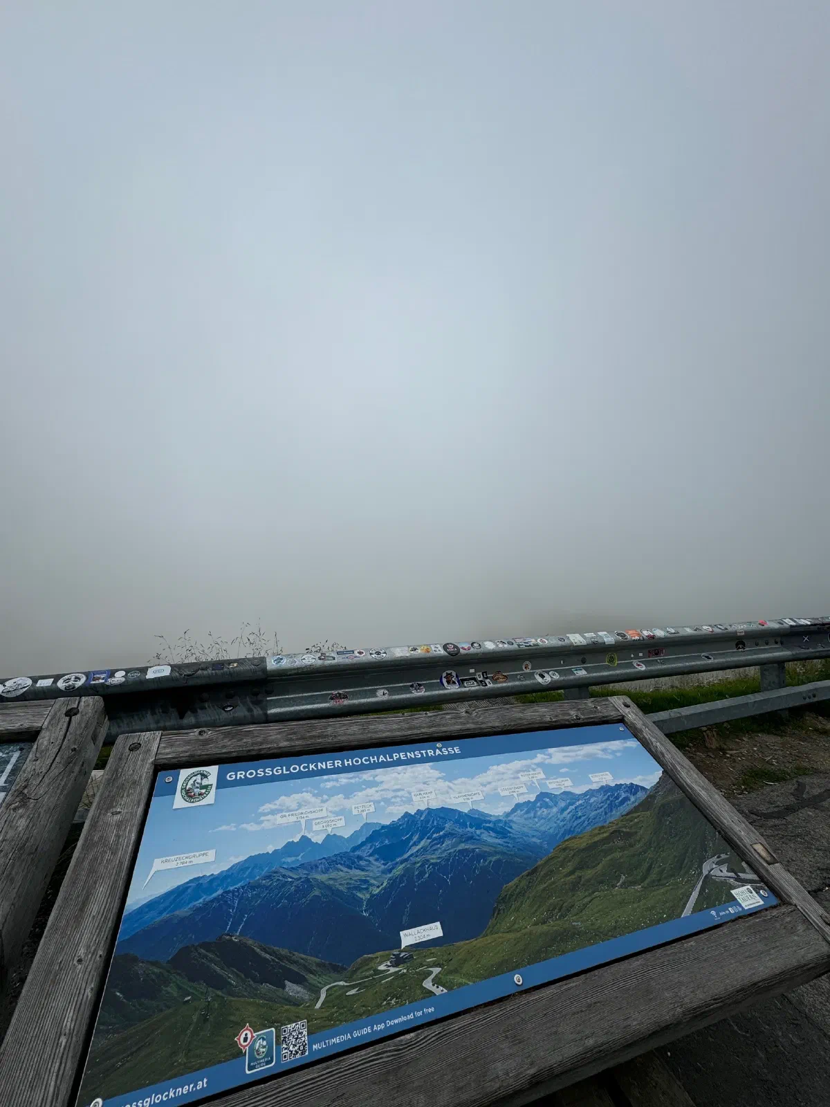 A panorama board of the Grossglockner High Alpine Road on a wooden frame, behind it a guardrail covered in stickers and a white wall of fog