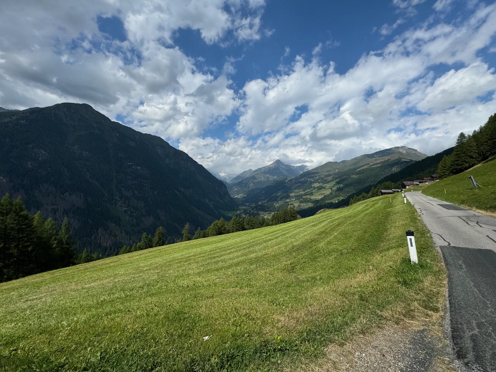 A narrow mountain road beside a mown meadow, behind it a wooded valley and a mountain range under a blue sky with clouds