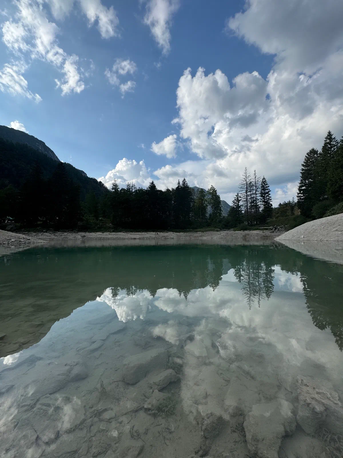 Clear, greenish shimmering water at the shore of Lake Predil, clouds and conifers reflected in it, wooded mountain slopes behind