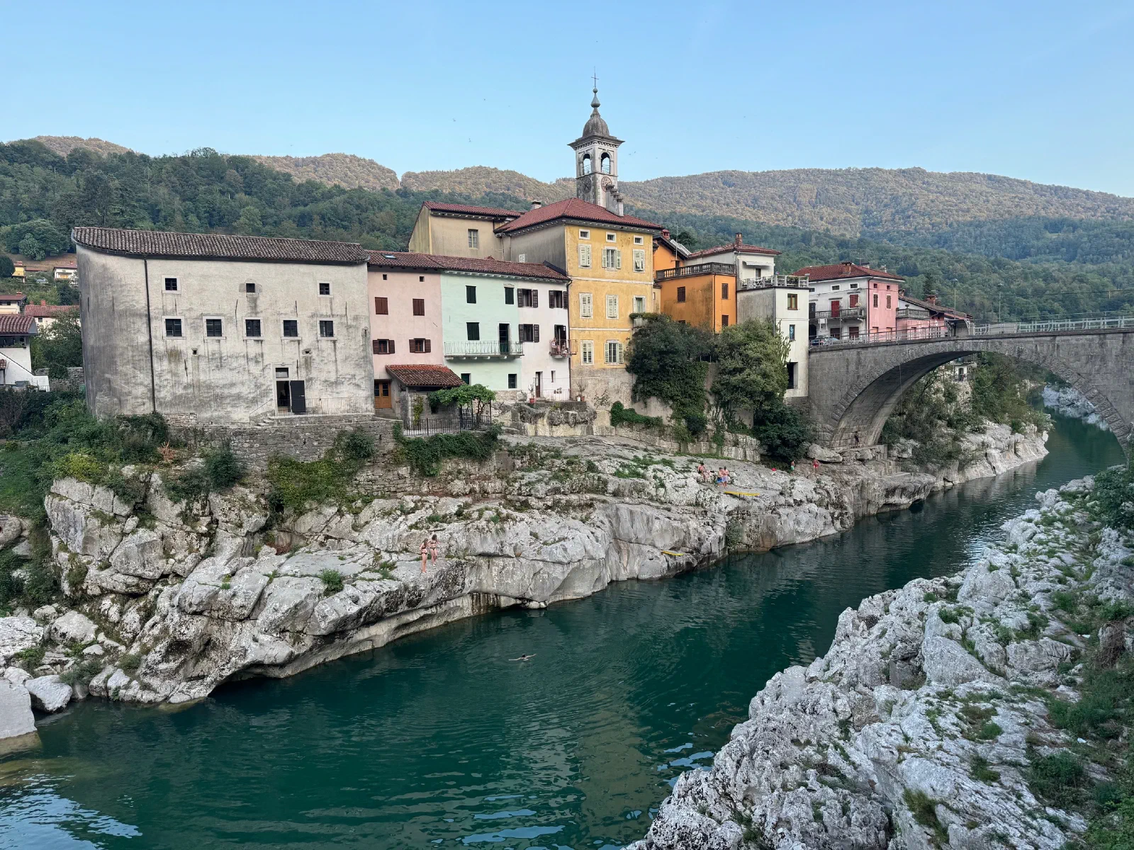 The old town of Kanal ob Soči with colorful houses, a church tower and a stone bridge over the turquoise Soča River, bright rocks in the foreground.