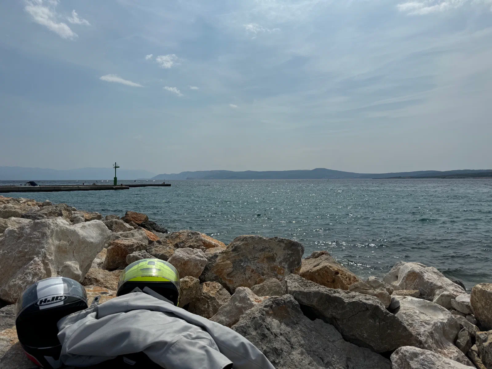 Two motorcycle helmets, one black and white and one neon yellow, and a gray motorcycle jacket lie on boulders at the shore, behind them a jetty with a small mast, the sea and low islands under a lightly clouded sky