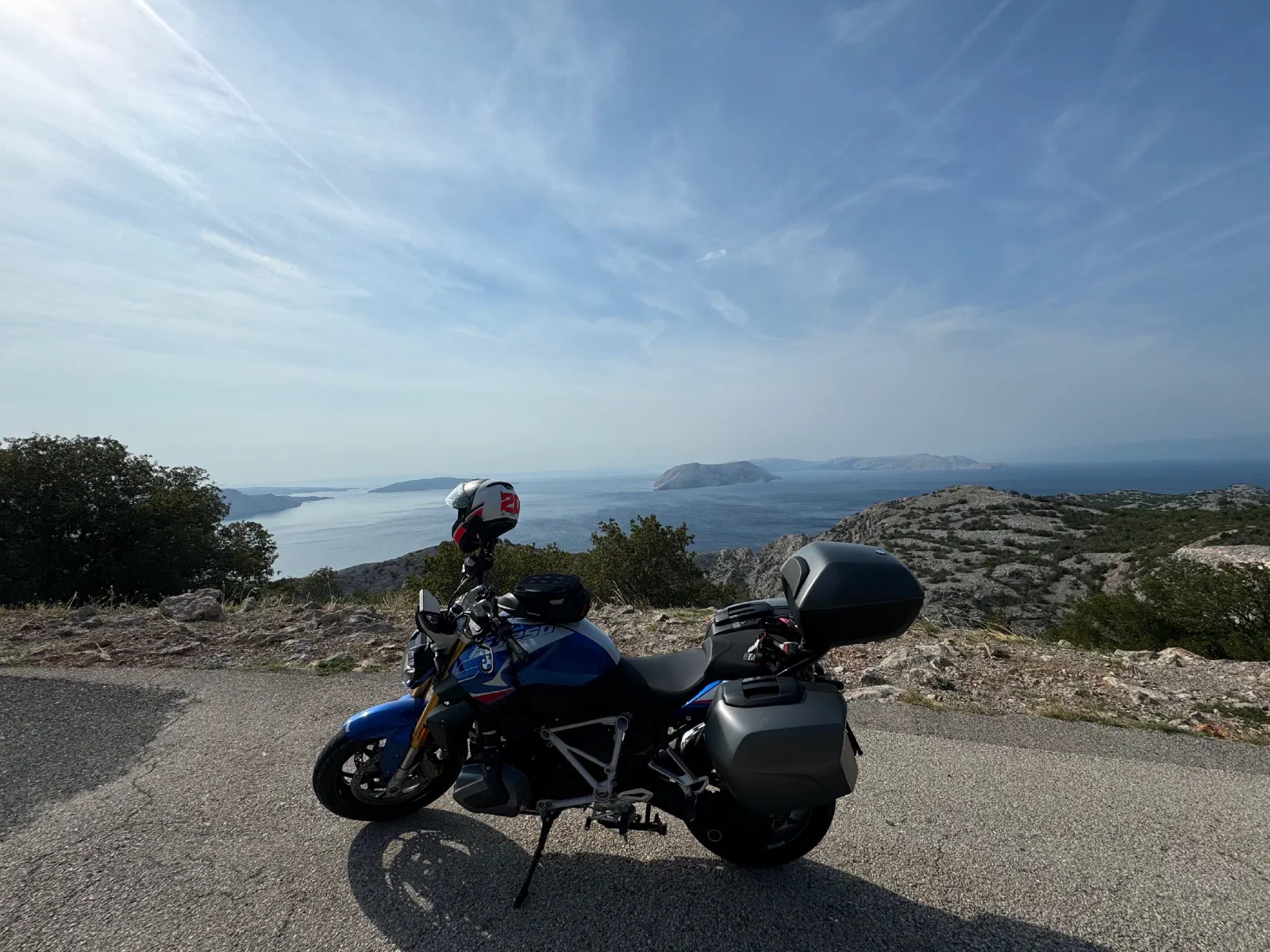 A blue-and-white motorcycle with side cases, a top case and a tank bag, seen from the side, stands on the asphalt at a rocky edge, behind it the sea with several islands under thin veils of cloud
