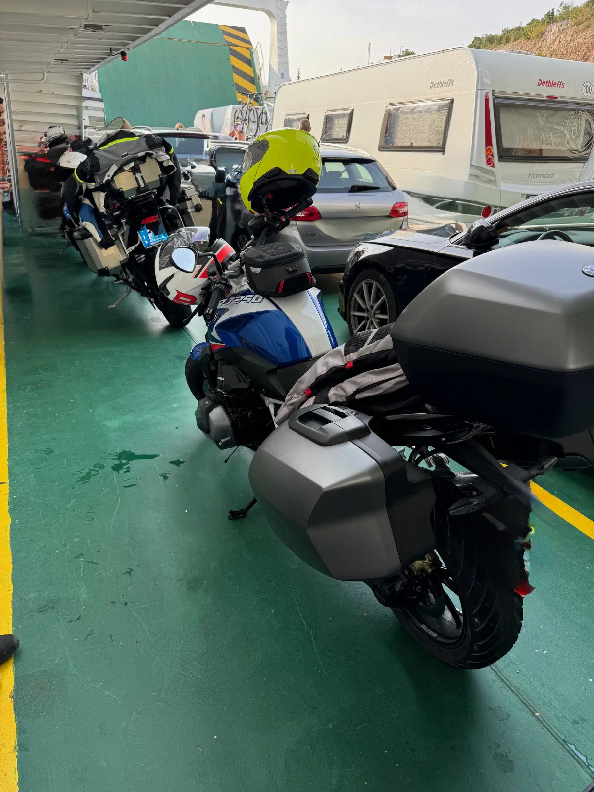 Car deck of a ferry with a green floor: a blue-and-white motorcycle with side cases, a top case and a neon-yellow helmet on the handlebars, in front of it another motorcycle with aluminum cases, next to it cars and a white travel trailer