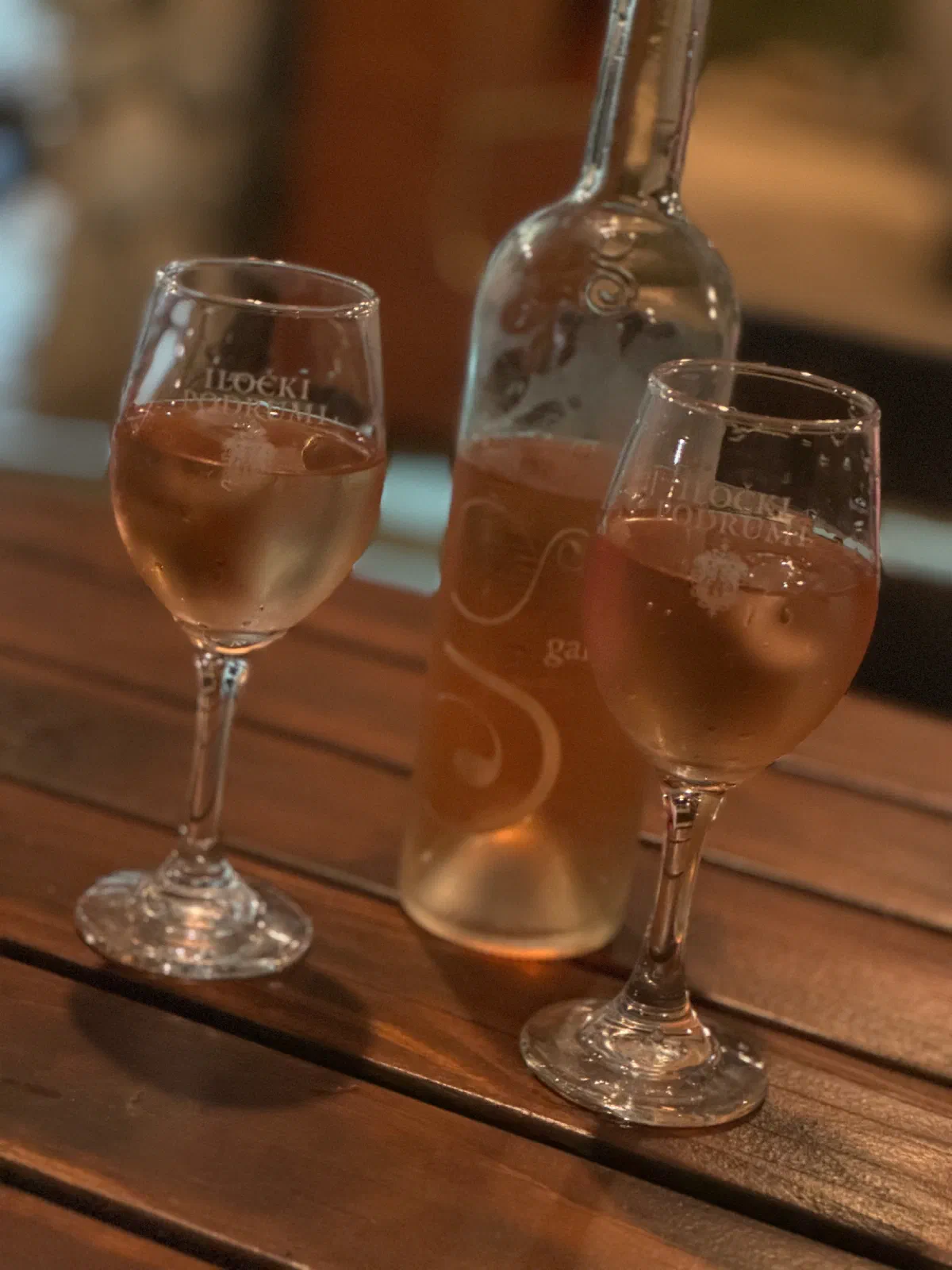 Two wine glasses with pale pink wine and a bottle stand on a dark wooden table, the background is blurred