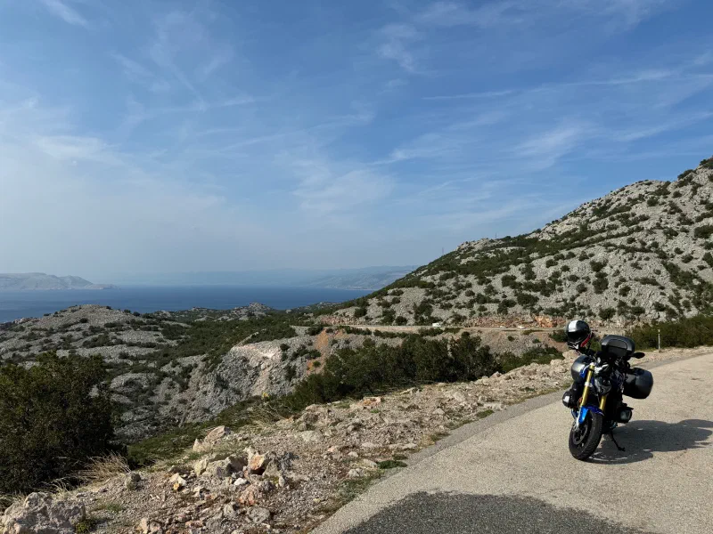 A blue-and-white motorcycle with side cases and a top case stands at the edge of a coastal road, behind it barren karst slopes with bushes and the sea under a blue sky with thin clouds.