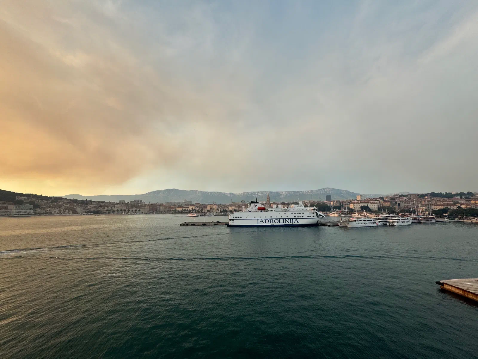 View across the harbor basin to Split: a white Jadrolinija ferry at the quay, behind it the old town with a tower and a mountain ridge, the sky hazy and tinged orange on the left