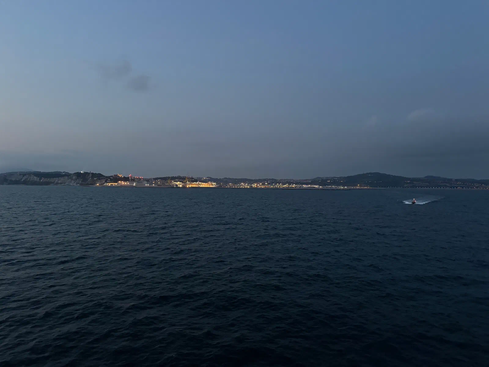Dark blue sea at dawn, on the horizon the lit coast with harbor facilities and a bright cliff, on the right a small boat with a wake