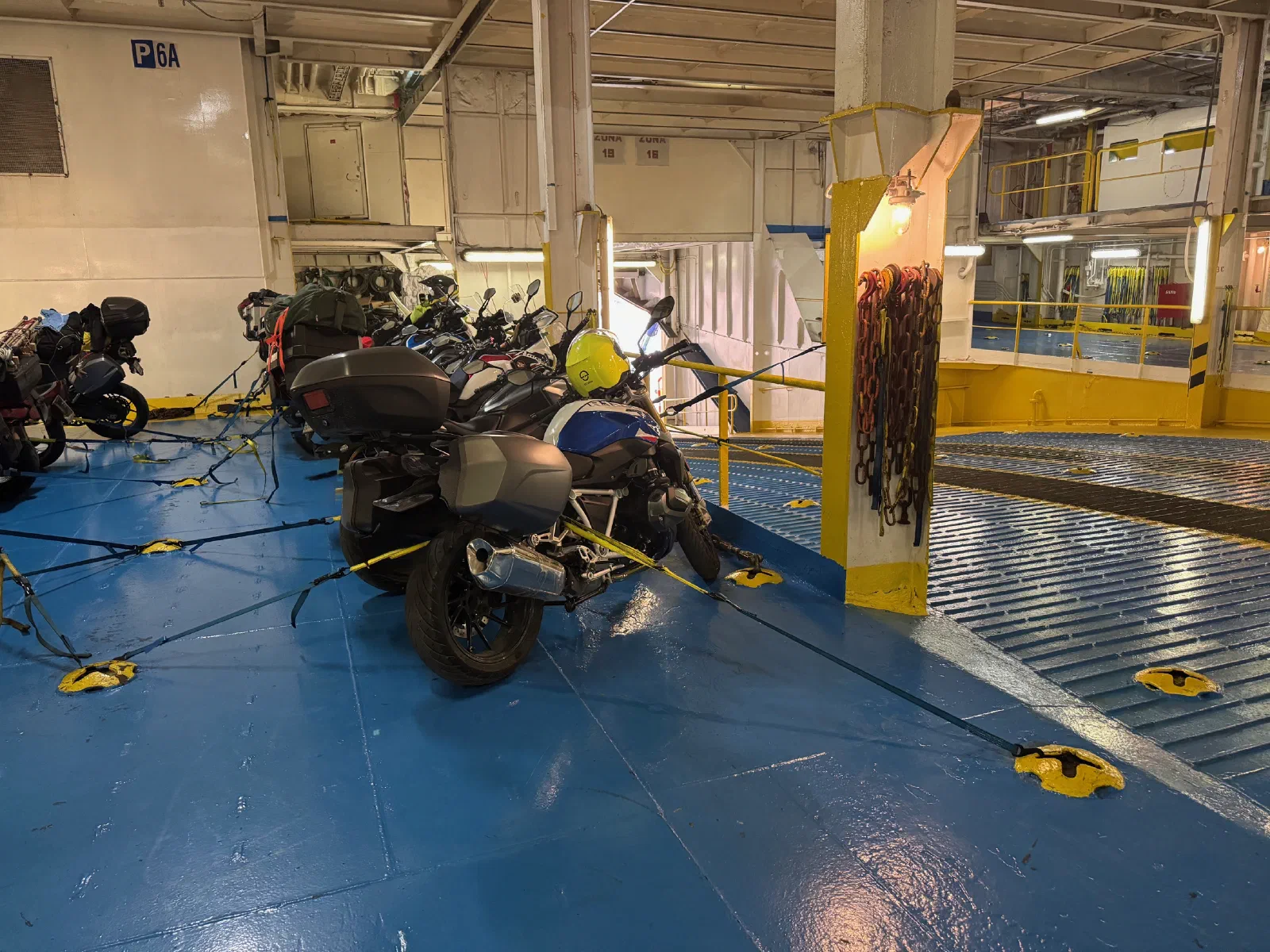 A motorcycle with side cases and a top case lashed down with yellow straps on the car deck of a ferry, a yellow helmet hanging from the mirror, more motorcycles behind it.