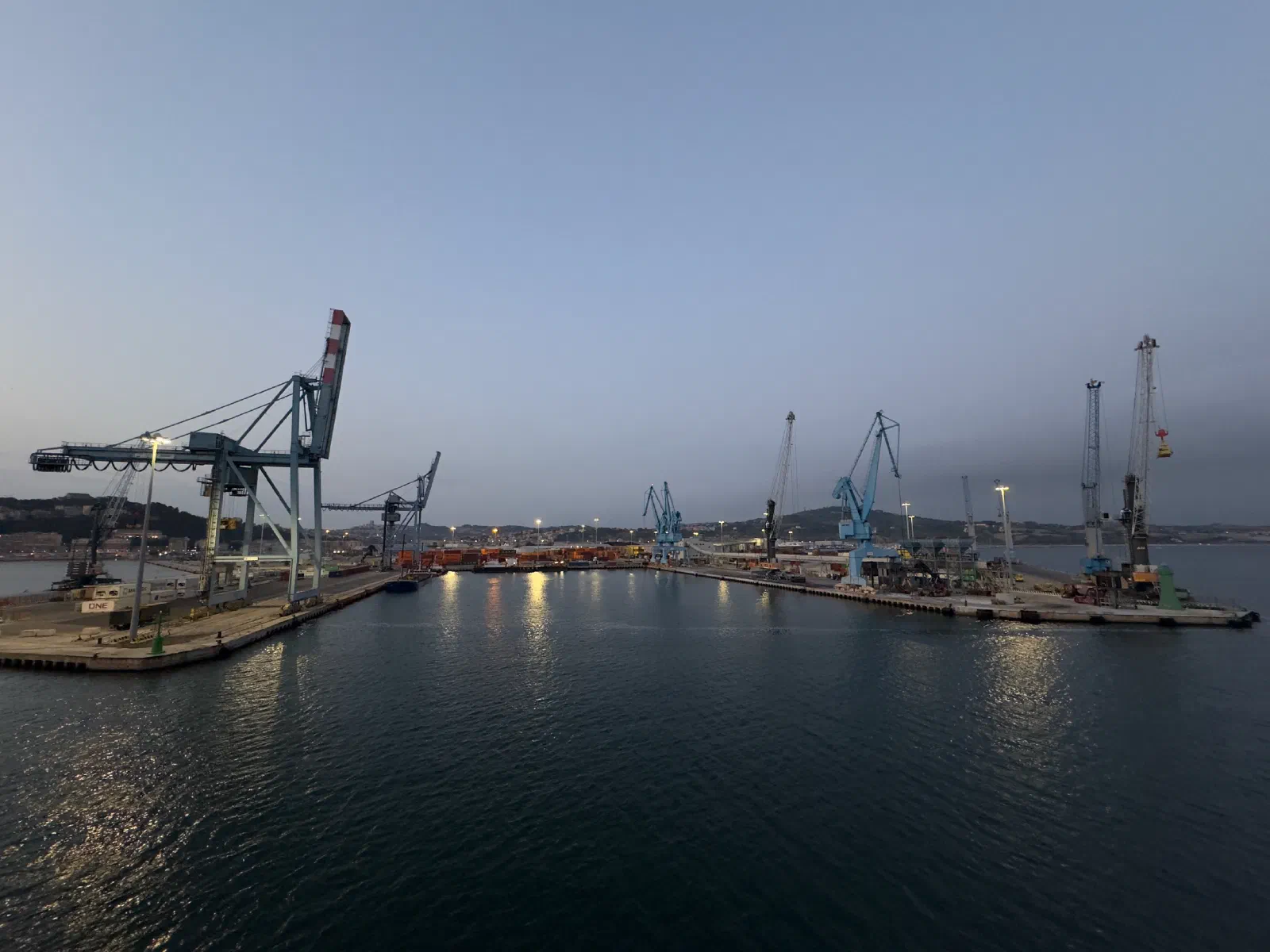 View from the ferry in the twilight across the calm harbor basin to container cranes and quays, behind them hills and the lights of the city