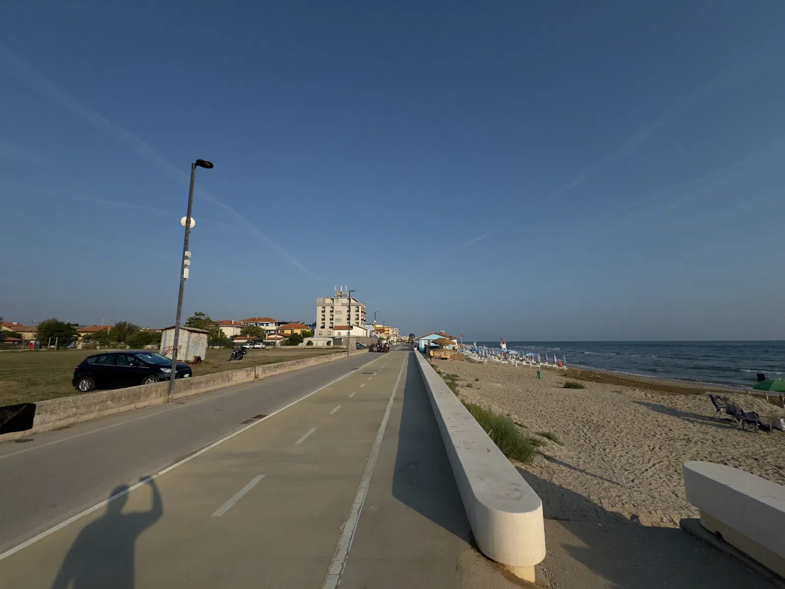 A road with a bike lane and a low concrete wall runs along the sandy beach, on the left a car on a lawn and a hotel building farther back, on the right beach and sea; in the lower left the photographer's shadow