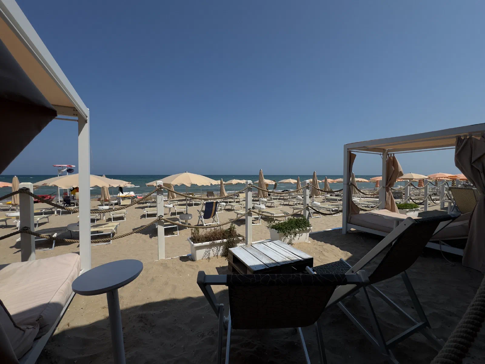 View between white posts with thick rope onto a stretch of beach with closed and open umbrellas and loungers, behind it the sea under a clear blue sky