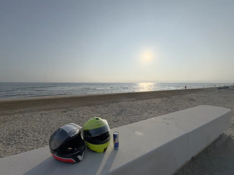 Two motorcycle helmets and a drink can sit on a concrete wall, behind them an almost empty sandy beach and the sea in the light of the low morning sun.