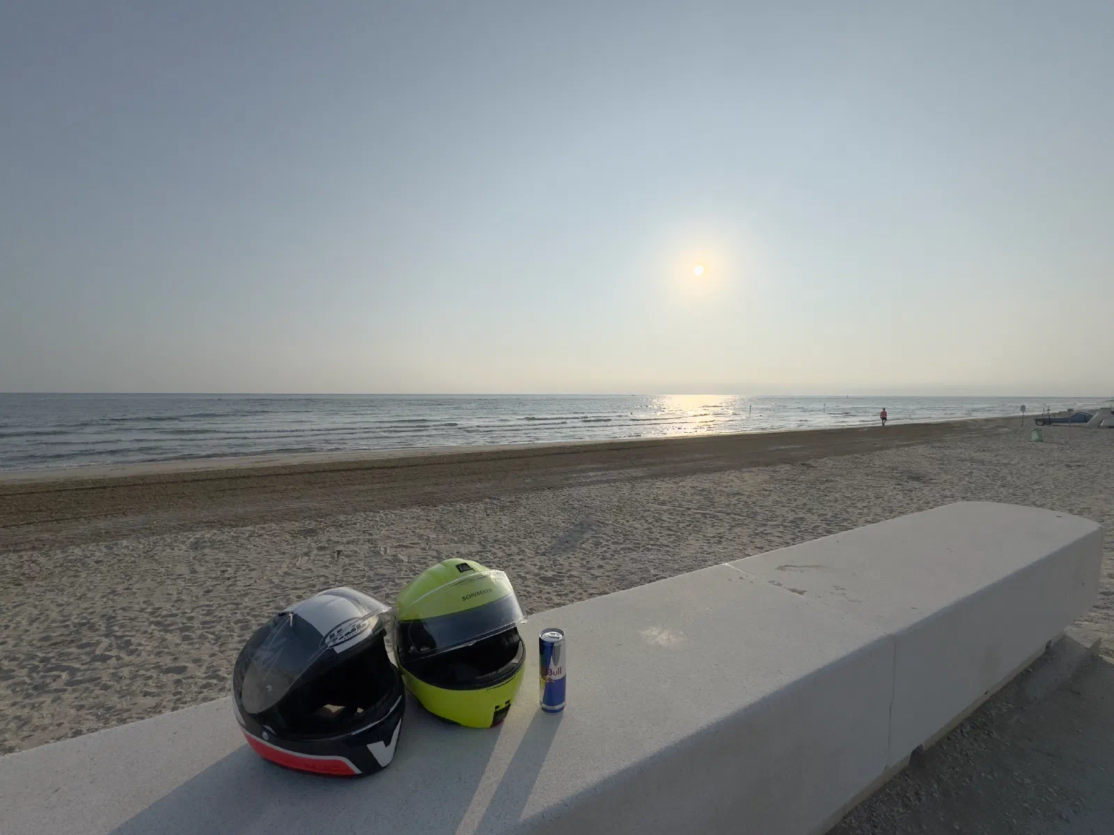 Two motorcycle helmets and a drink can sit on a concrete wall, behind them an almost empty sandy beach and the sea in the light of the low morning sun.