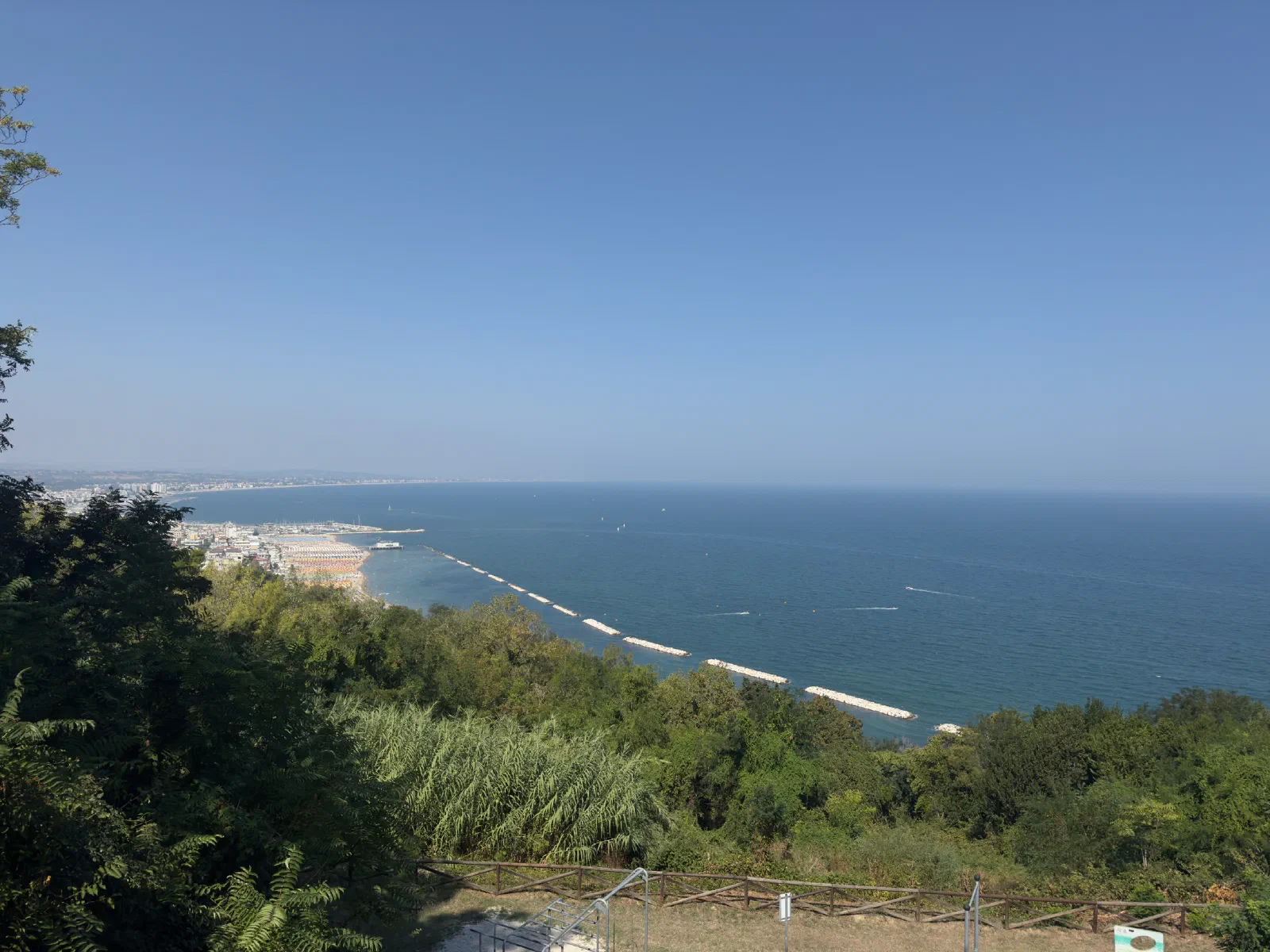 View from a hill over a wooded slope to a beach and the blue sea, in the water a row of light-colored blocks parallel to the coast