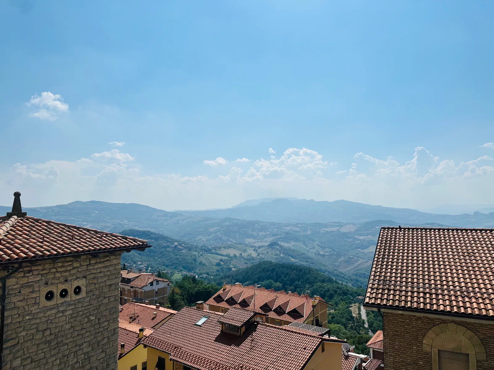 View over tile roofs and stone houses to wooded hills and distant mountain ranges under a light blue sky with a few clouds