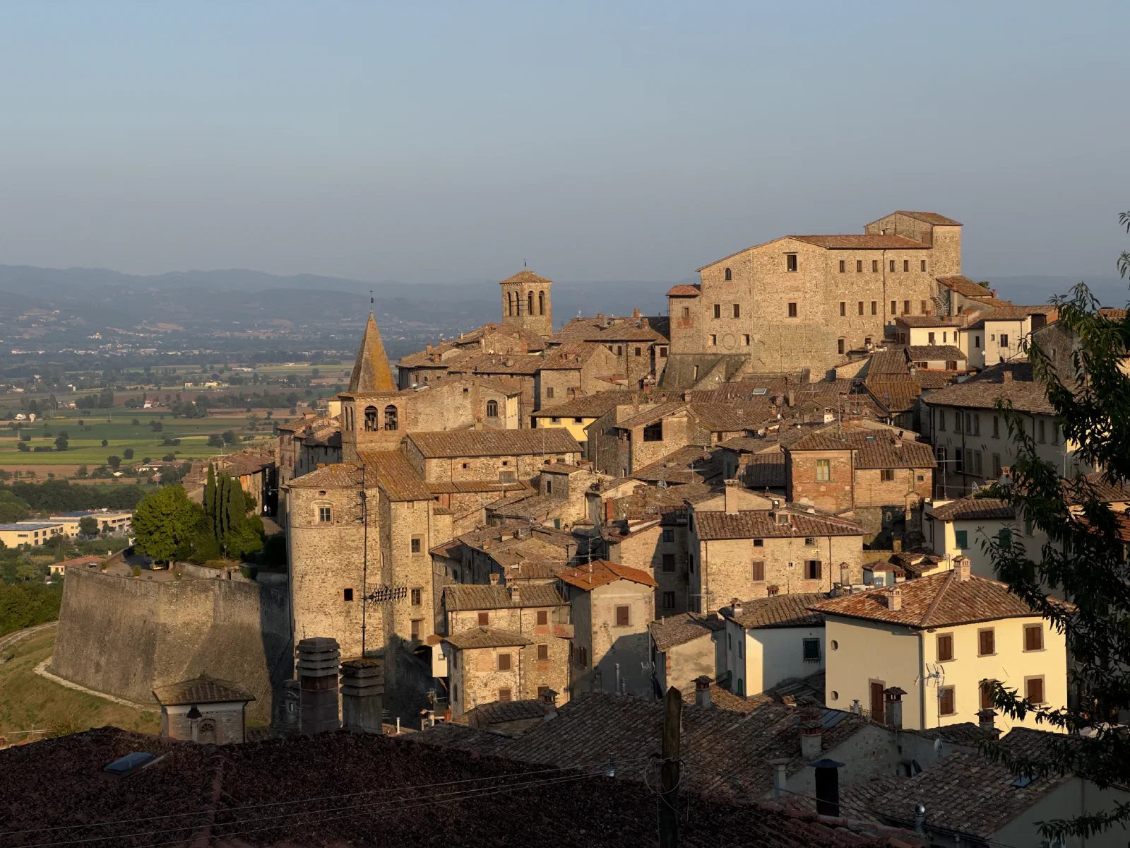 Roofs and towers of the medieval old town of Anghiari in warm evening light, behind it a wide valley in the haze