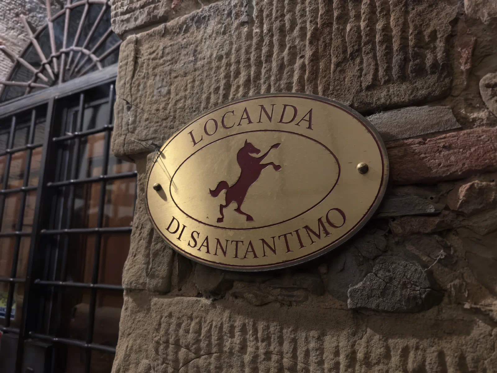 An oval brass sign reading Locanda di Santantimo with a rearing horse on a sandstone wall