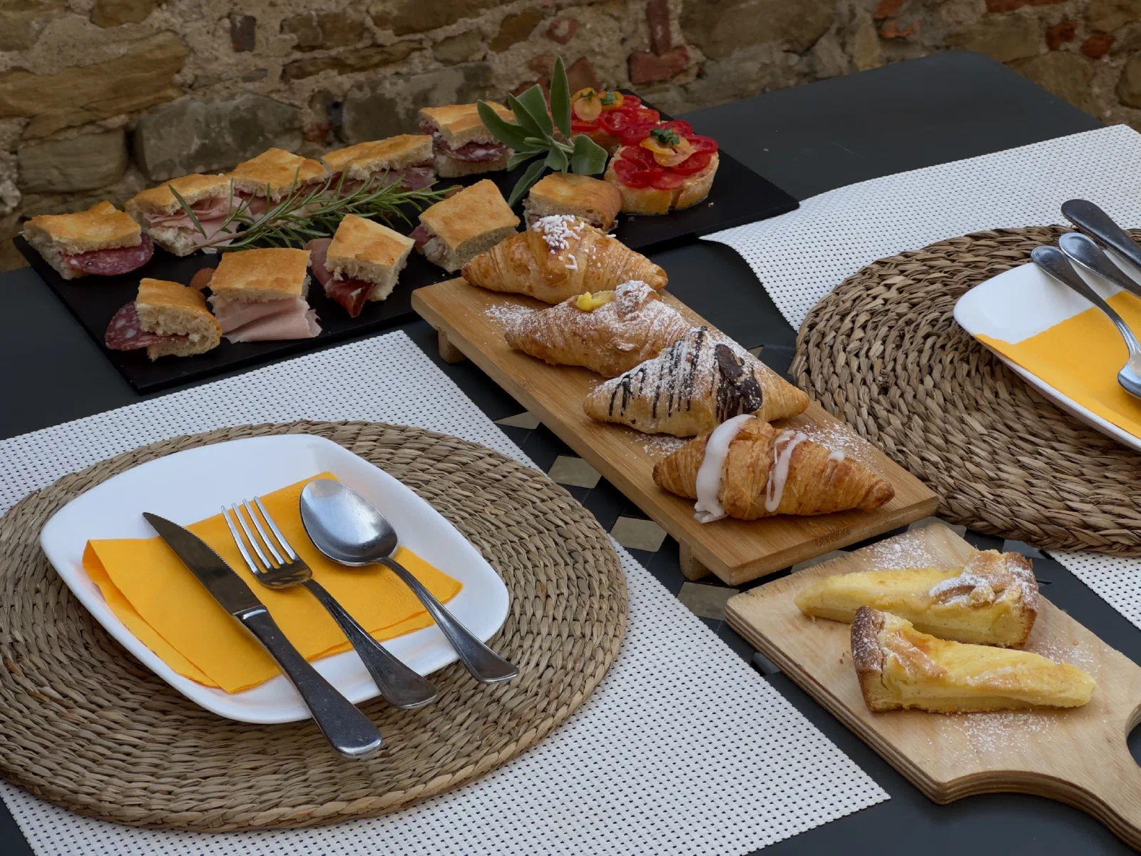A set breakfast table with two place settings, behind it a platter of topped slices of bread, croissants and pastries in front of a stone wall