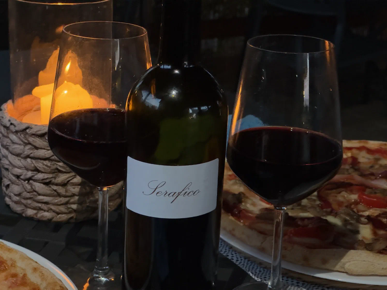 Two glasses of red wine and a bottle of wine next to a candle in a glass lantern, in front of them pizza on plates, on a dark table in the evening.