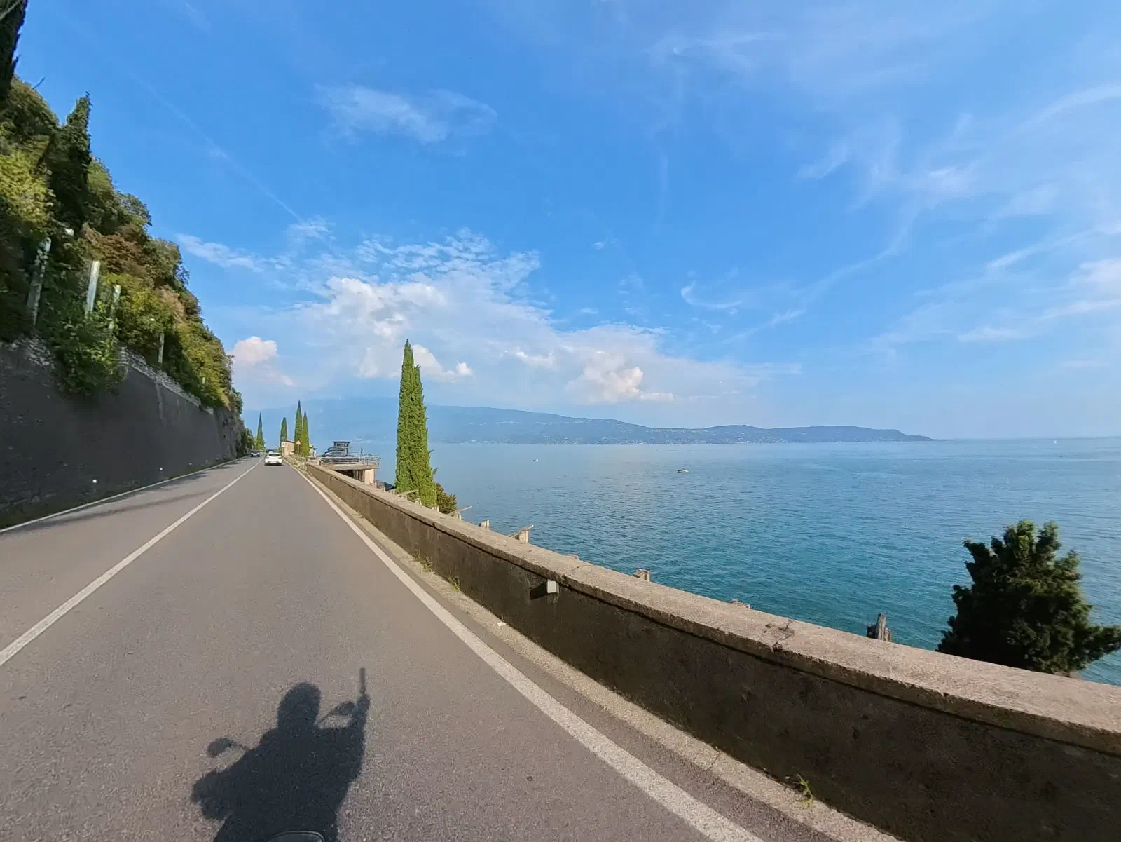 A lakeside road runs straight ahead; on the right, behind a parapet wall, Lake Garda stretches to the far shore, with a slender cypress in between; on the asphalt, the shadow of the rider with a raised hand