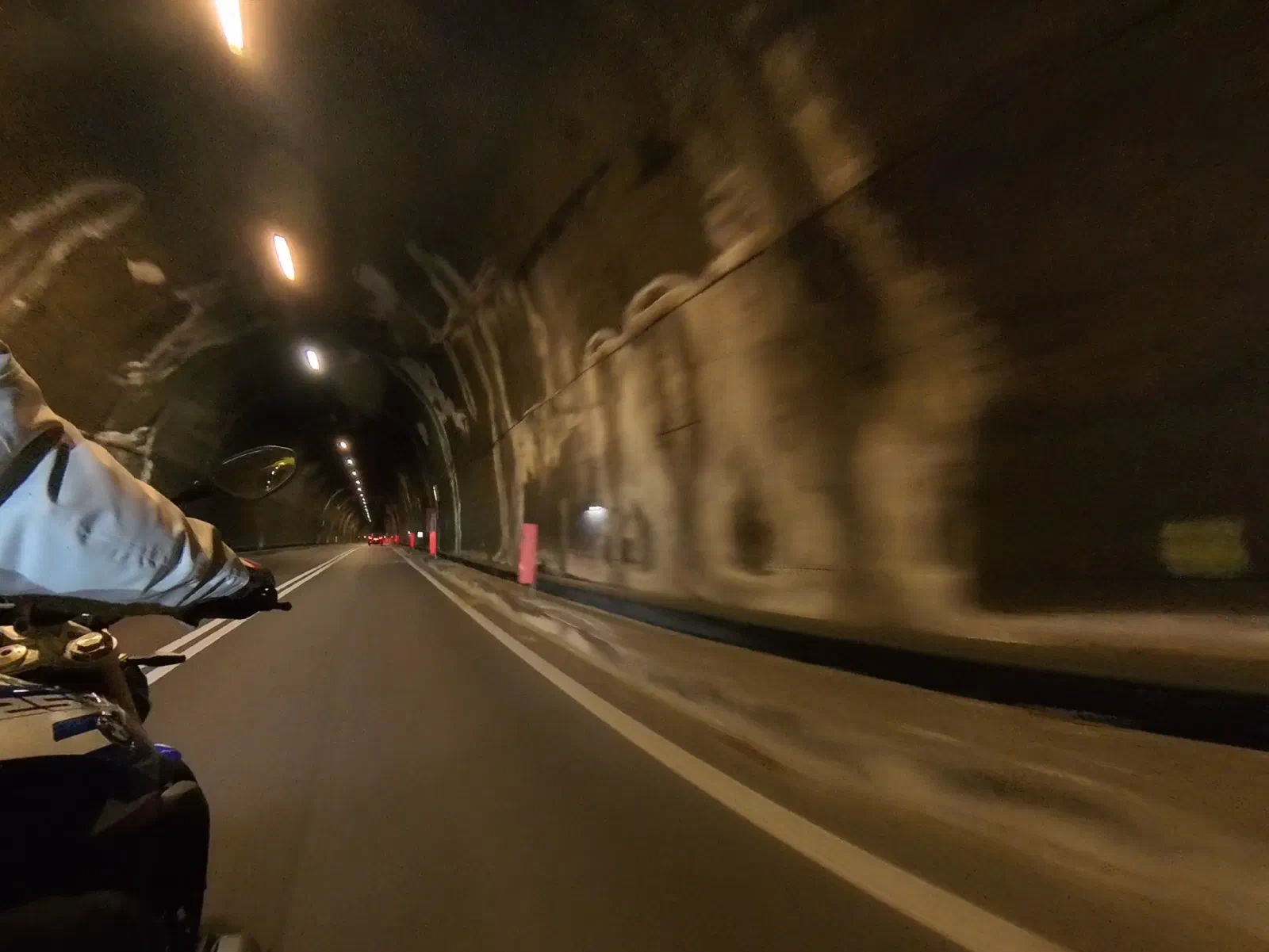 A lit road tunnel with a rough, unlined rock wall; in the left foreground the rider's arm and glove