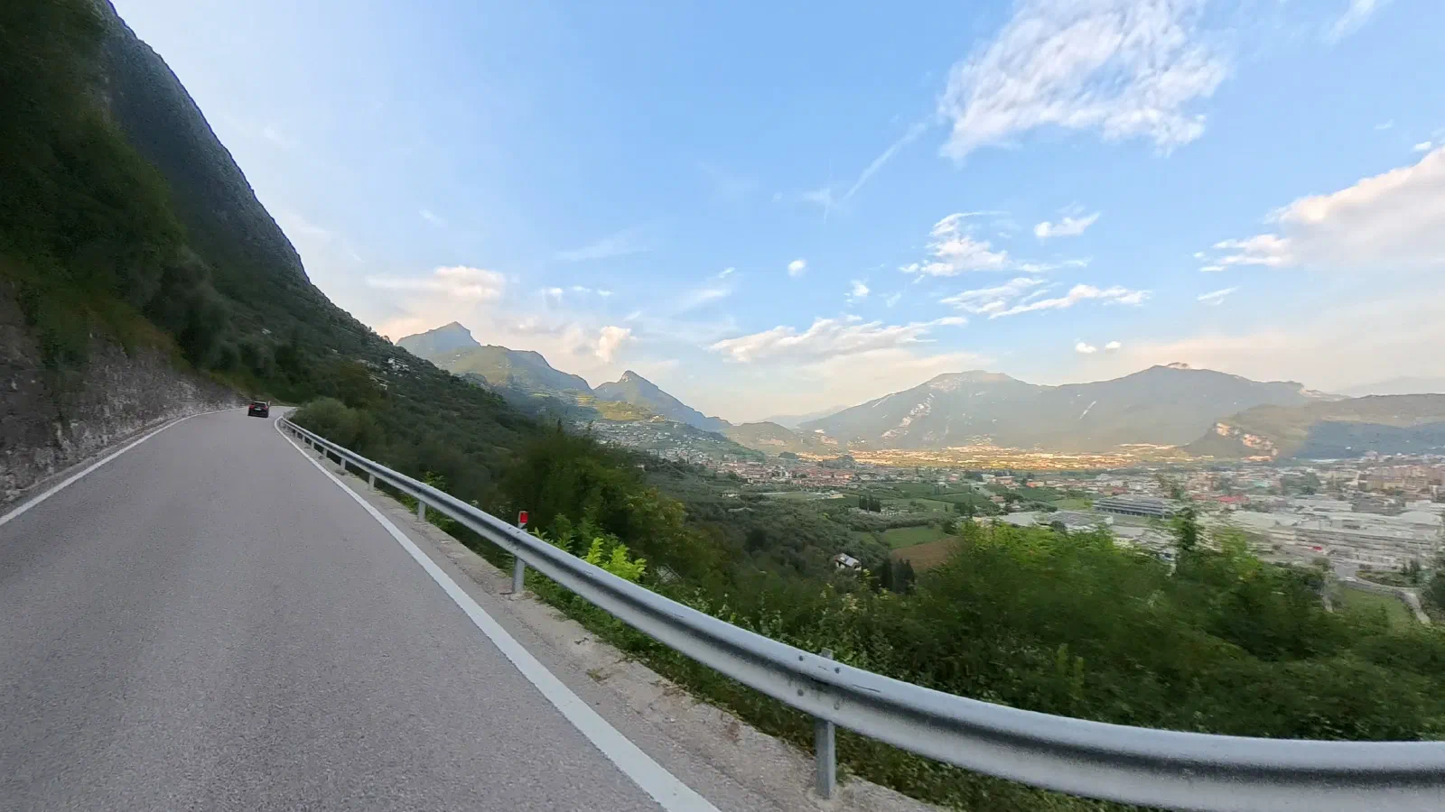 A mountain road with a guardrail runs along the hillside, below it a wide valley with towns, behind it mountain ranges under light clouds