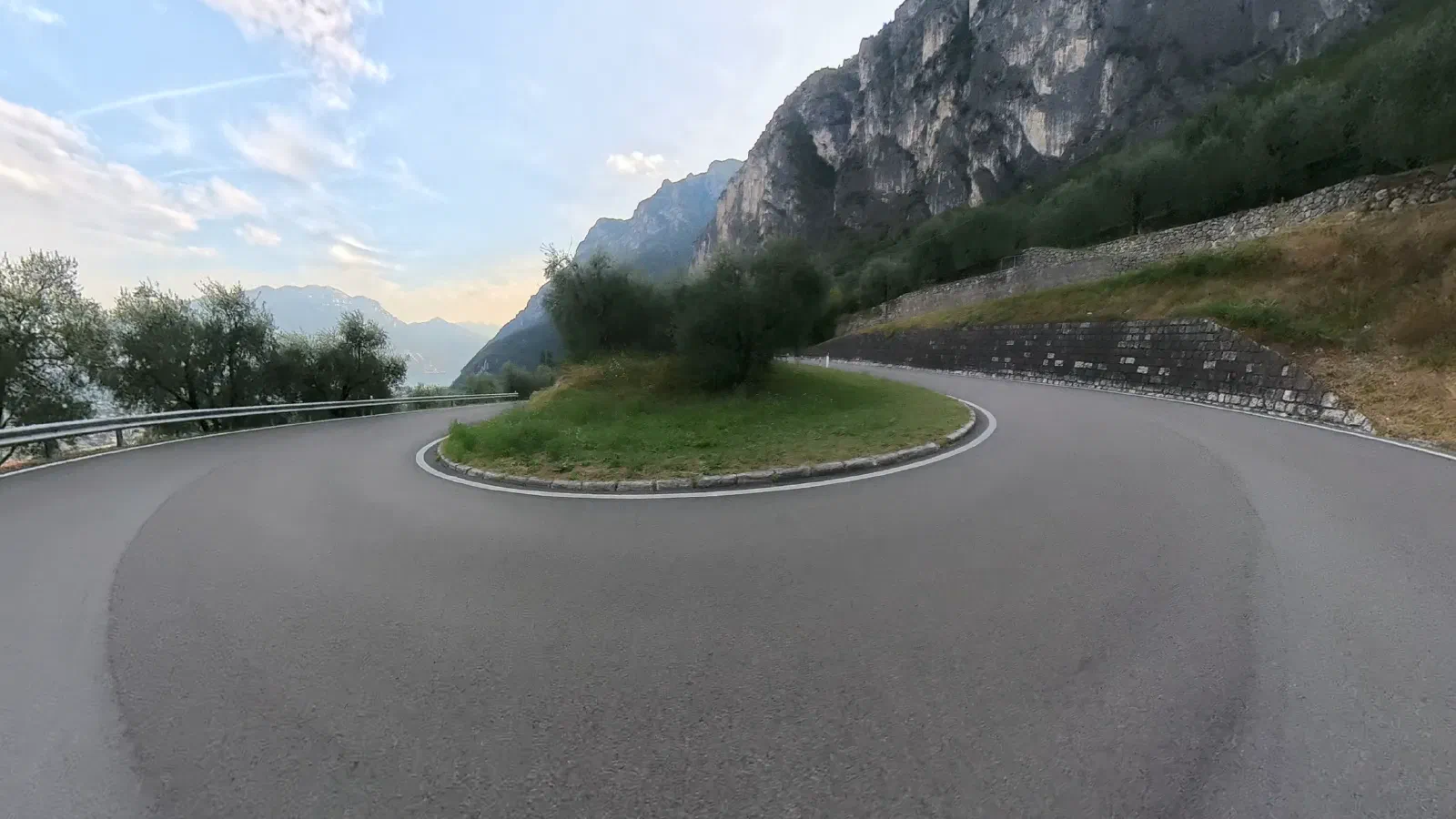 A hairpin bend around a grassy island with an olive tree, above it a tall, light-colored rock face