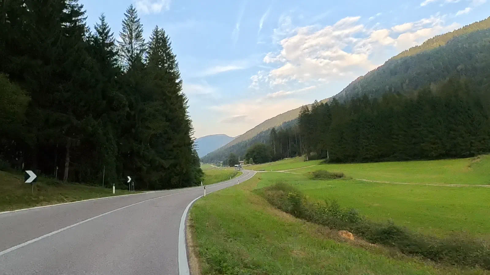 A country road between a green meadow and a spruce forest, a bus far ahead, behind it wooded mountain slopes in the evening light