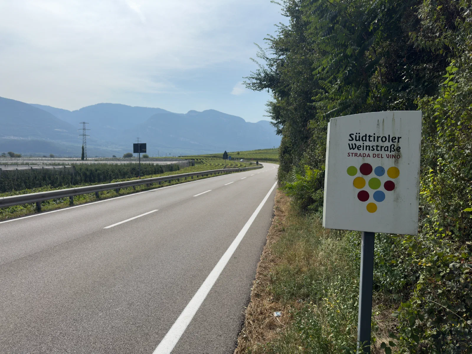 An empty road with a guardrail winds through the valley, on the right the sign Südtiroler Weinstraße with colorful dots, on the left orchards, behind them mountain ranges in the haze