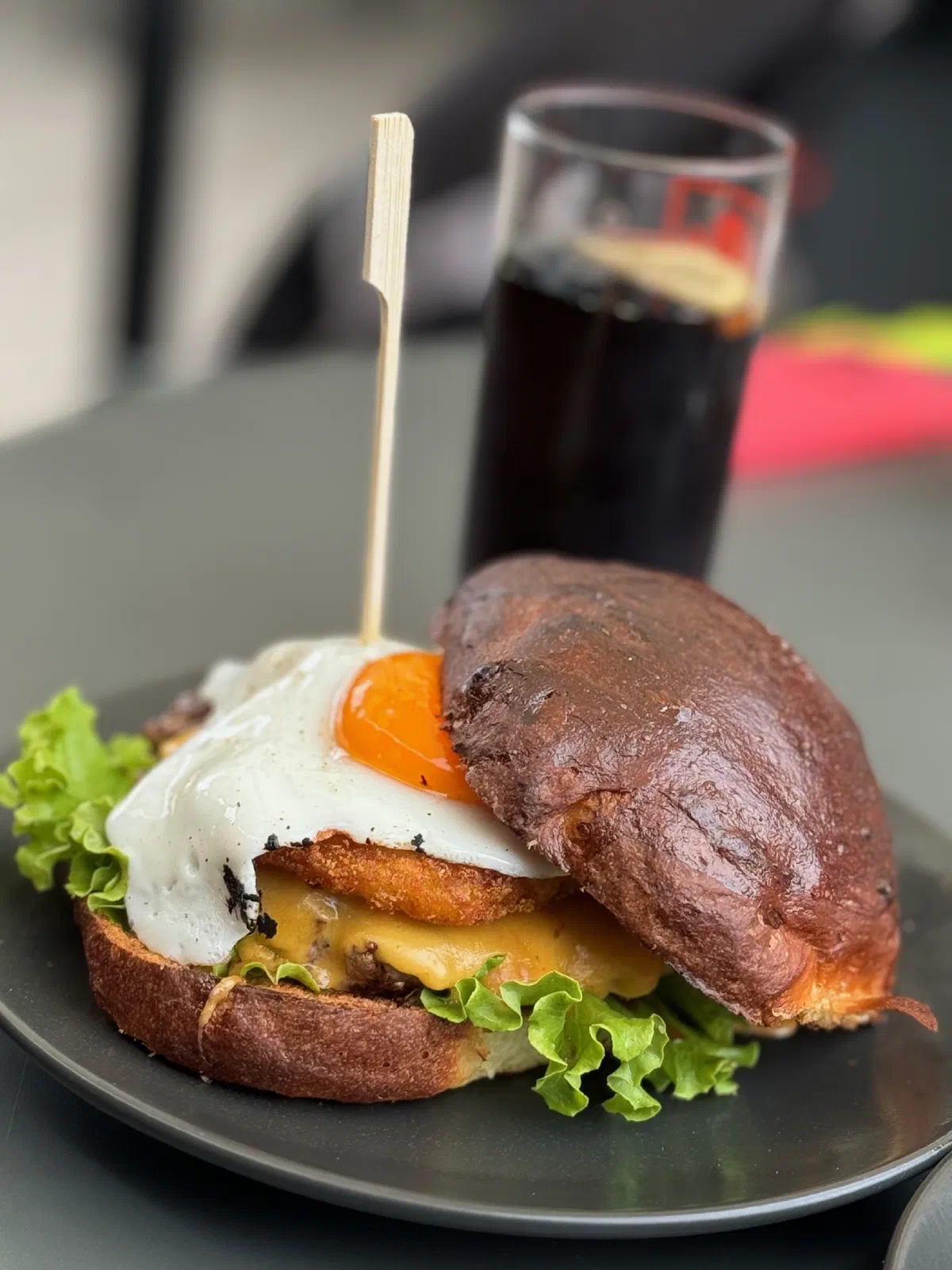 A burger with a fried egg, cheese and lettuce in a dark-baked bun, a wooden skewer stuck in it, on a dark plate; behind it, blurred, a glass with a dark drink