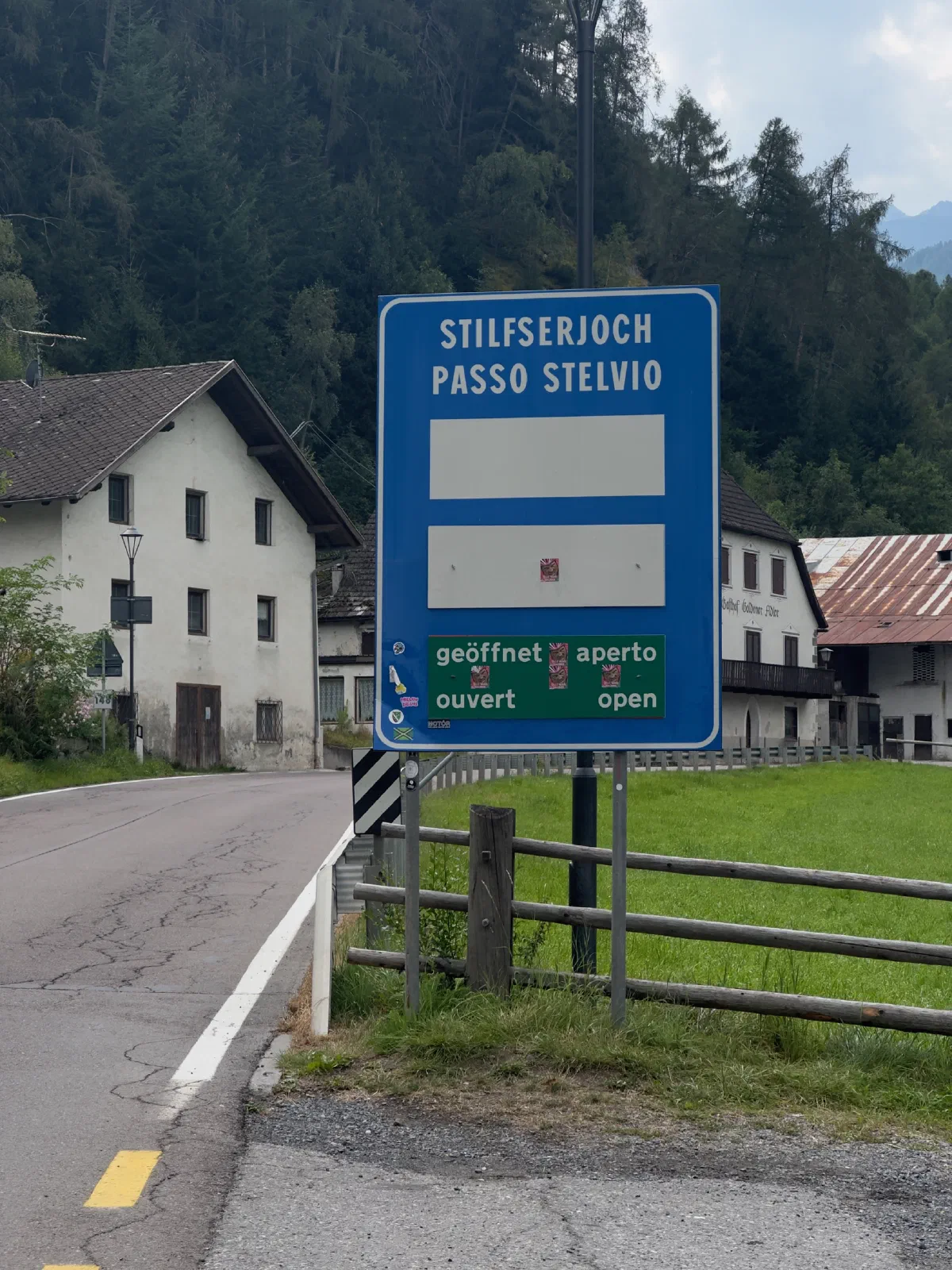 A blue sign reading Stilfserjoch, Passo Stelvio with a green panel saying geöffnet, aperto, ouvert, open stands beside the road, behind it old stone houses and conifer forest