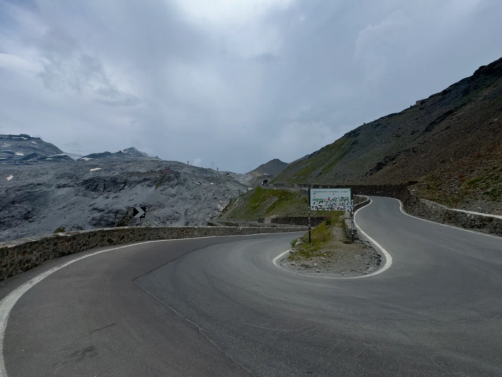 A hairpin of the Stelvio Pass road under a gray cloudy sky, in the bend a board with many stickers, on the left gray rock with masts, on the right a steep scree slope; no vehicle on the road
