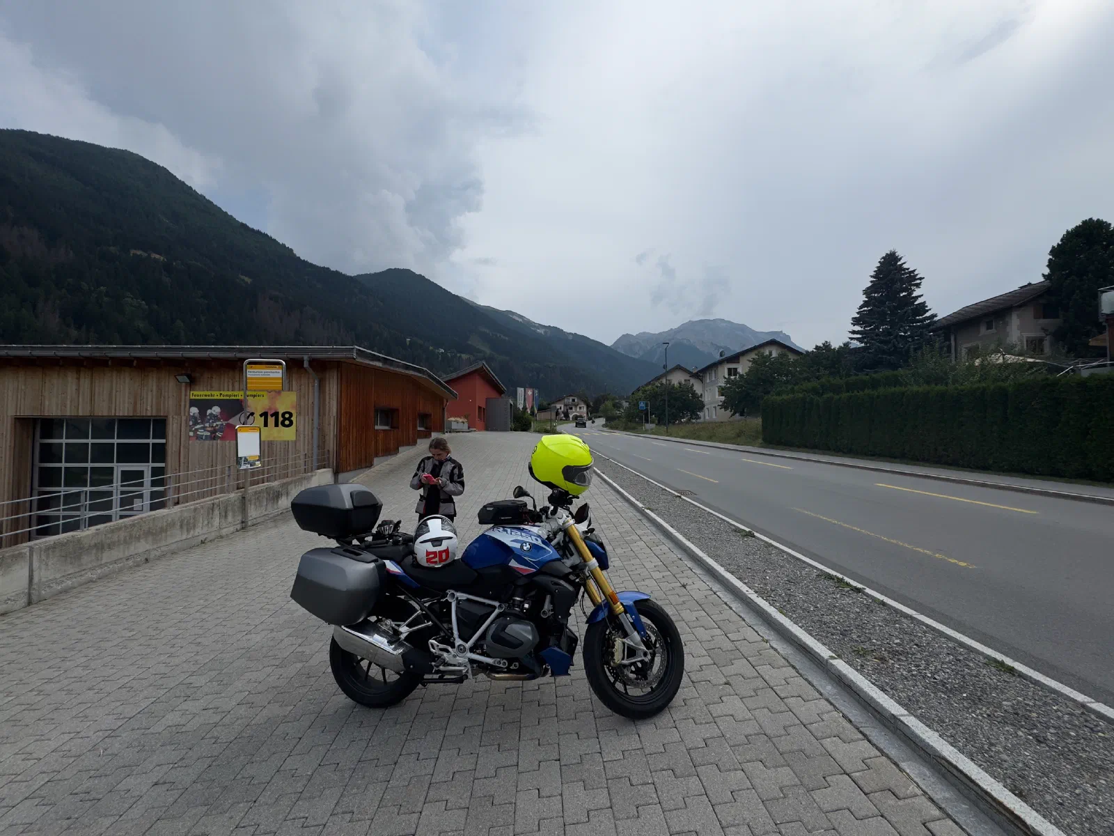 The motorcycle with side cases and a top case stands on a paved square next to a wooden building, a yellow helmet on the handlebars, a white helmet on the seat, behind it a person in a riding suit, on the right the road in front of wooded mountains under dark clouds