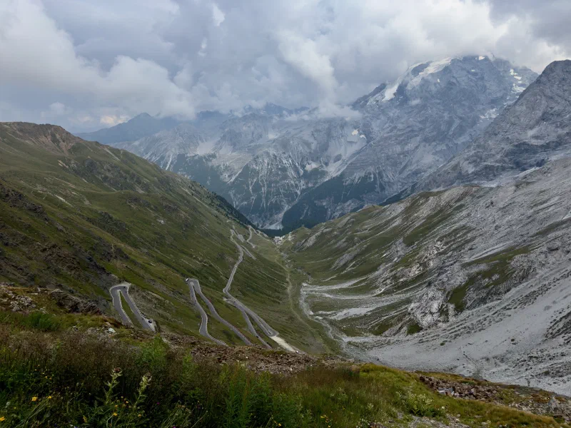 View from the lookout plateau just below the pass summit down onto the hairpins of the Stelvio Pass road, behind them mountains with glacier remnants in thick clouds.